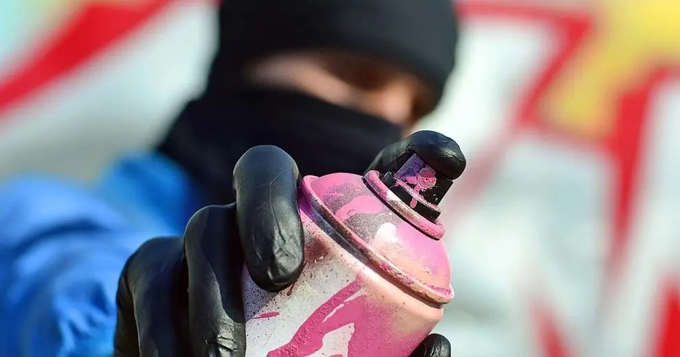 A young graffiti artist in a blue jacket and black mask is holding a can of paint in front of him against a background of colored graffiti drawing. Street art and vandalism concept.