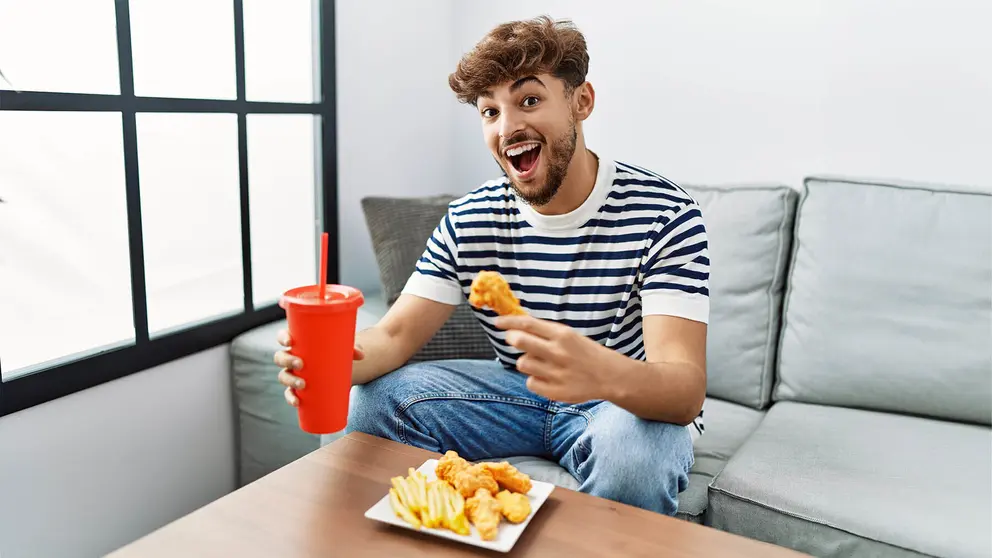 Joven comiendo pollo frito en su sof&aacute; sobre una mesa elevable