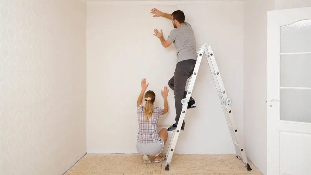 Un hombre subido en una escalera de metal y una mujer ayudan a aplicar papel pintado en la pared de la habitaci&oacute;n