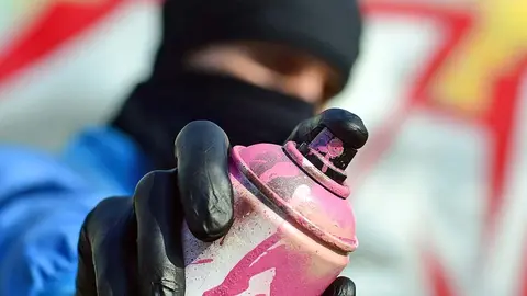 A young graffiti artist in a blue jacket and black mask is holding a can of paint in front of him against a background of colored graffiti drawing. Street art and vandalism concept.