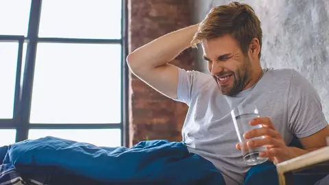 young man with headache holding glass of water in bed at home