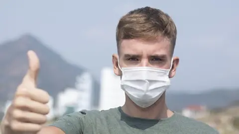 Serious European man, young handsome guy in protective sterile medical mask on face looking at camera show thumb up, like gesture. Air pollution, virus, Chinese pandemic coronavirus protection concept