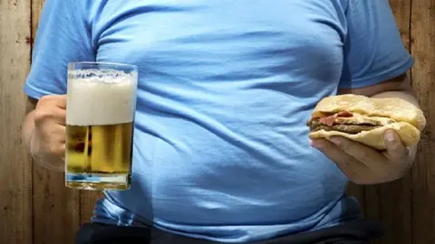 Fat man with beers and hamburger on his hand over wooden wall background